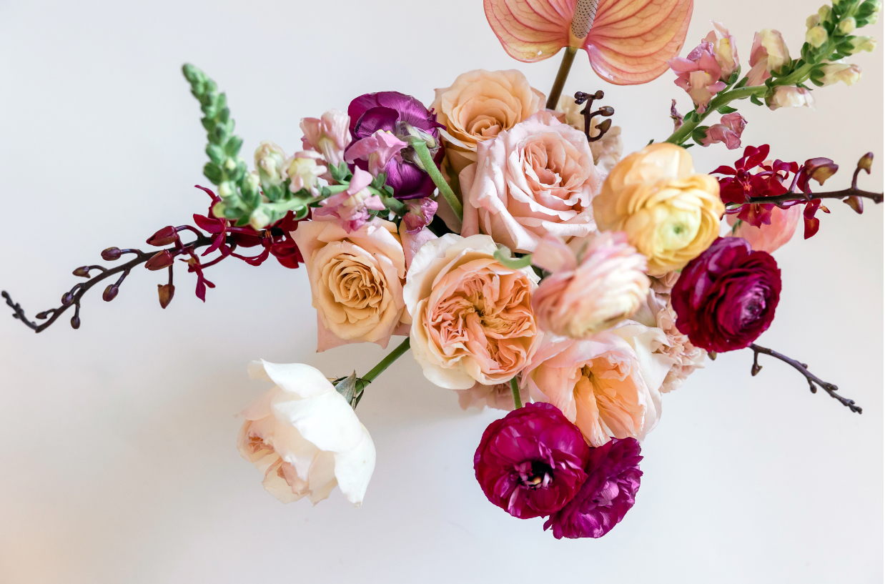 Elegant mixed flower bouquet with roses, ranunculus, snapdragons, and anthurium in soft and bold tones on white background.