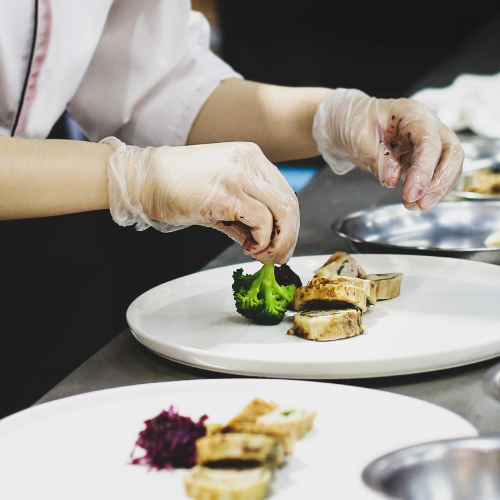 A chef wearing gloves carefully plating a gourmet meal with broccoli and sliced meat in a professional kitchen.