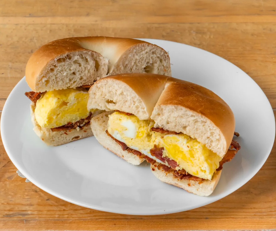 Two freshly made breakfast bagel sandwiches served on a white plate, filled with fluffy eggs and crispy bacon on soft, golden-brown bagels, placed on a wooden table.